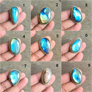 May include: A collection of nine gemstone rings, each featuring a different iridescent labradorite stone set in a gold-colored metal band. The stones vary in shape, including oval, teardrop, and irregular forms, displaying blue, green, and yellow hues.