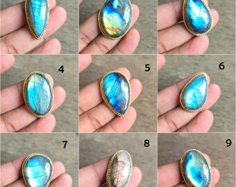 May include: A collection of nine gemstone rings, each featuring a different iridescent labradorite stone set in a gold-colored metal band. The stones vary in shape, including oval, teardrop, and irregular forms, displaying blue, green, and yellow hues.