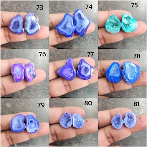 May include: A collection of paired, polished agate geodes in various shades of blue, purple, and green. Each geode has a unique crystalline interior and a smooth, rounded exterior. The geodes are displayed on a hand.