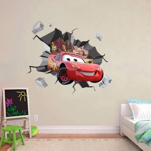 May include: A red Lightning McQueen car wall sticker breaking through a wall, with a Route 66 sign in the background. The sticker has a 3D effect with grey rock and debris.
