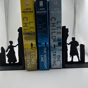 Puede incluir: Sujeta libros de metal negro con la silueta de dos figuras de pie en un acantilado rocoso. Los sujeta libros sostienen tres libros de bolsillo de Lee Child, titulados "Die Trying", "The Midnight Line" y "Echo Burning".