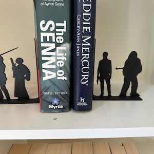 Peut inclure: Des serre-livres en métal noir avec des silhouettes de personnages à côté de deux livres. Les livres sont intitulés "The Life of Senna" et "Eddie Mercury". Les serre-livres sont sur une étagère blanche.