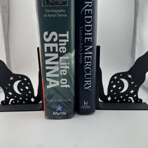 Puede incluir: Dos sujetalibros de metal negro con forma de gatos con un diseño de luna creciente y estrellas. Los sujetalibros sostienen dos libros, uno titulado "The Life of Senna" y el otro titulado "Freddie Mercury".