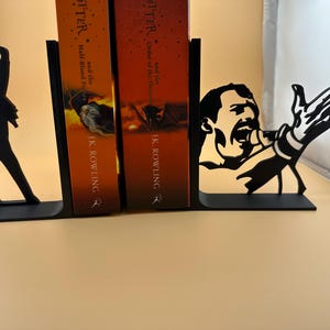 K&ouml;nnte beinhalten: Schwarze Metall-Buchst&uuml;tzen mit Silhouetten eines S&auml;ngers in ikonischen Posen. Eine Buchst&uuml;tze zeigt eine Figur mit erhobenem Arm, die andere einen S&auml;nger mit Mikrofon. Die Buchst&uuml;tzen sind zum Halten von B&uuml;chern konzipiert.