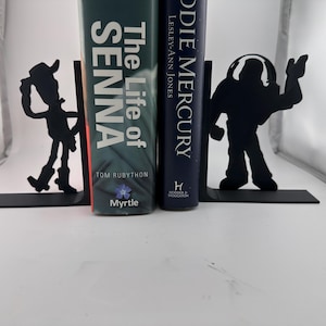 May include: Black metal bookends with cowboy and space ranger silhouette designs. The bookends support two books, "The Life of Senna" and "Oddie Mercury". The bookends are on a white surface.