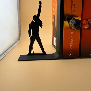K&ouml;nnte beinhalten: Schwarzer Metall-Buchst&uuml;tze mit einer Silhouette einer Person mit erhobenem Arm. Die Buchst&uuml;tze befindet sich neben einem Stapel B&uuml;cher mit orangefarbenen Einb&auml;nden. Die Buchst&uuml;tze dient dazu, B&uuml;cher aufrecht zu halten.