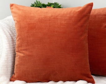 Op de afbeelding: Twee vierkante, oranje kussens met een getextureerde, corduroy-achtige uitstraling. De kussens liggen op een witte, zachte deken en een grijze bank. De kussens geven de ruimte een warme, uitnodigende uitstraling.