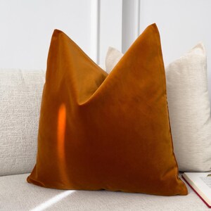 Puede incluir: Un gran cojín cuadrado de terciopelo naranja quemado es el punto focal, apoyado en un sofá de color claro. Un segundo cojín más pequeño, blanquecino, está detrás. La escena sugiere un ambiente acogedor de sala de estar, con un libro y una taza visibles.