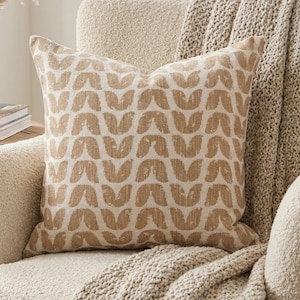 May include: A decorative throw pillow with a repeating pattern of tan-colored shapes on a cream background. The pillow is placed on a cream-colored armchair with a matching throw blanket. A vase with dried plants sits nearby.
