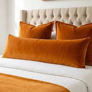 May include: A set of burnt orange velvet bedding consisting of two square throw pillows, one long rectangular lumbar pillow, and a matching bed runner. The items are arranged on a white quilted bedspread against a tufted beige headboard.