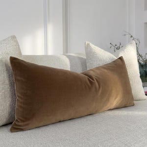 May include: A rectangular lumbar cushion made of smooth brown velvet fabric positioned in front of two square off-white textured throw cushions. The cushions are arranged on a light grey upholstered sofa surface with a soft, consistent finish.