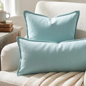 May include: Two rectangular throw cushions in a solid light blue colour with a visible woven fabric texture. The cushions feature a flange edge detail and are positioned on a textured off-white upholstered chair with a cream-coloured knit throw.