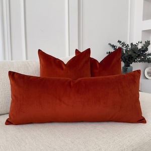 May include: A long rectangular lumbar pillow and two square pillows, all crafted from a solid burnt orange velvet fabric. The pillows feature a smooth, soft-touch texture and are arranged together on a light-colored, textured fabric sofa.