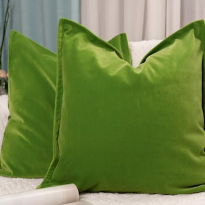 May include: Two green velvet throw pillows on a white couch. The pillows are square and have a smooth, soft texture.