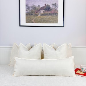 May include: A white linen couch with five white linen pillows, including a long lumbar pillow. The couch is in front of a white wall with a framed picture of a cottage in a field.