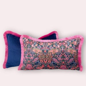 May include: Two rectangular cushions. One is navy blue with a pink fringe. The other features a peacock and floral pattern on a dark blue background, also with a pink fringe.