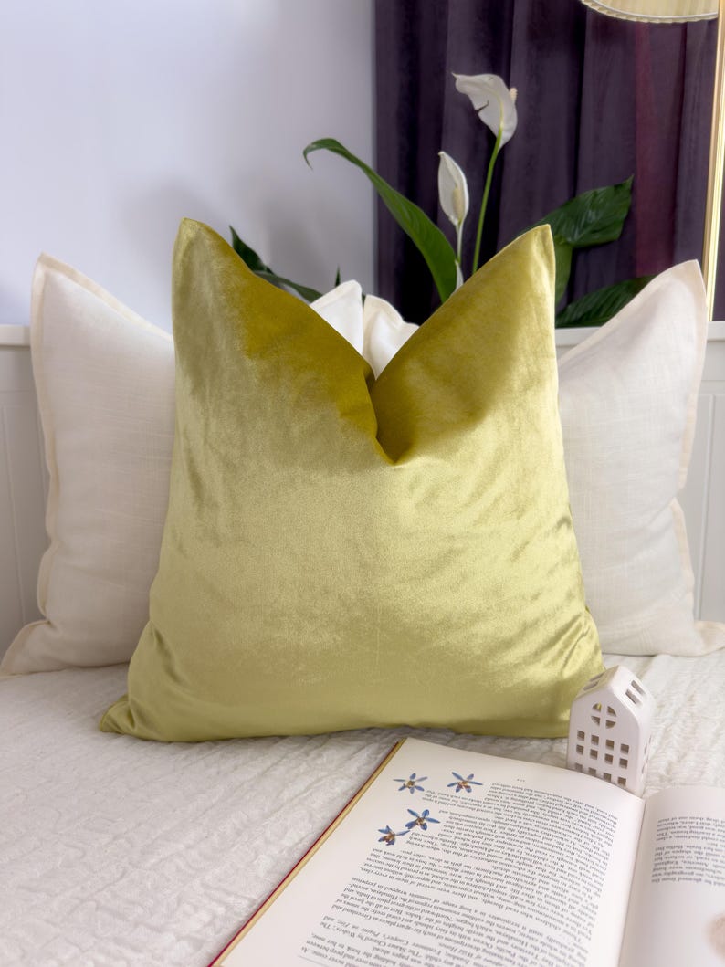 K&ouml;nnte beinhalten: Eine dekorative Anordnung mit drei Kissen. Ein gro&szlig;es, chartreusefarbenes Samtkissen steht im Mittelpunkt, flankiert von zwei cremefarbenen Kissen. Ein offenes Buch mit floralen Illustrationen und eine kleine wei&szlig;e Hausfigur sind im Vordergrund.