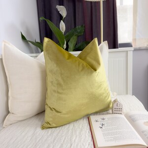 K&ouml;nnte beinhalten: Ein Bett mit zwei Kissen, eines cremefarben und eines aus chartreusefarbenem Samt. Ein offenes Buch mit rotem Einband liegt auf dem Bett, und eine kleine wei&szlig;e hausf&ouml;rmige Dekoration befindet sich in der N&auml;he. Eine Lampe mit cremefarbenem Schirm ist im Hintergrund.