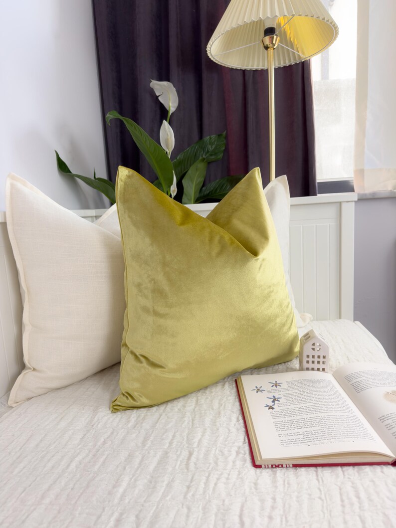K&ouml;nnte beinhalten: Ein Bett mit zwei Kissen, eines cremefarben und eines aus chartreusefarbenem Samt. Ein offenes Buch mit rotem Einband liegt auf dem Bett, und eine kleine wei&szlig;e hausf&ouml;rmige Dekoration befindet sich in der N&auml;he. Eine Lampe mit cremefarbenem Schirm ist im Hintergrund.