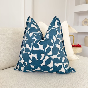 May include: A teal blue throw pillow with a white floral pattern. The pillow is sitting on a white couch.