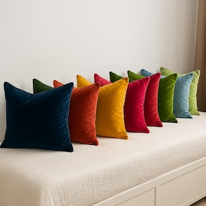 May include: A row of colorful velvet throw pillows in shades of navy, orange, yellow, red, green, and blue. The pillows are arranged on a white upholstered bench, creating a vibrant and inviting display.