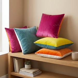 Könnte beinhalten: Eine Sammlung farbenfroher Samt-Zierkissen in Fuchsia, Türkis, Gelb und Orange. Die Kissen sind auf einem Holzregal angeordnet, einige gestapelt und andere angelehnt. Die Kissen haben eine weiße oder orangefarbene Rückseite. Maße nicht sichtbar.
