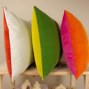 May include: Three decorative throw pillows with contrasting colours. The pillow on the left is white with a red border. The middle pillow is green with a yellow border. The pillow on the right is orange with a pink border.