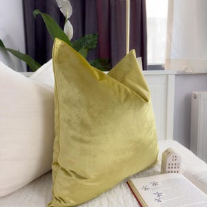 K&ouml;nnte beinhalten: Ein kissenf&ouml;rmiges Dekokissen aus chartreusefarbenem Samt liegt auf einem Bett neben einem wei&szlig;en Kissen. Das Kissen hat ein weiches, pl&uuml;schiges Aussehen. Ein Buch mit rotem Buchr&uuml;cken und ein kleines dekoratives Haus liegen ebenfalls auf dem Bett.