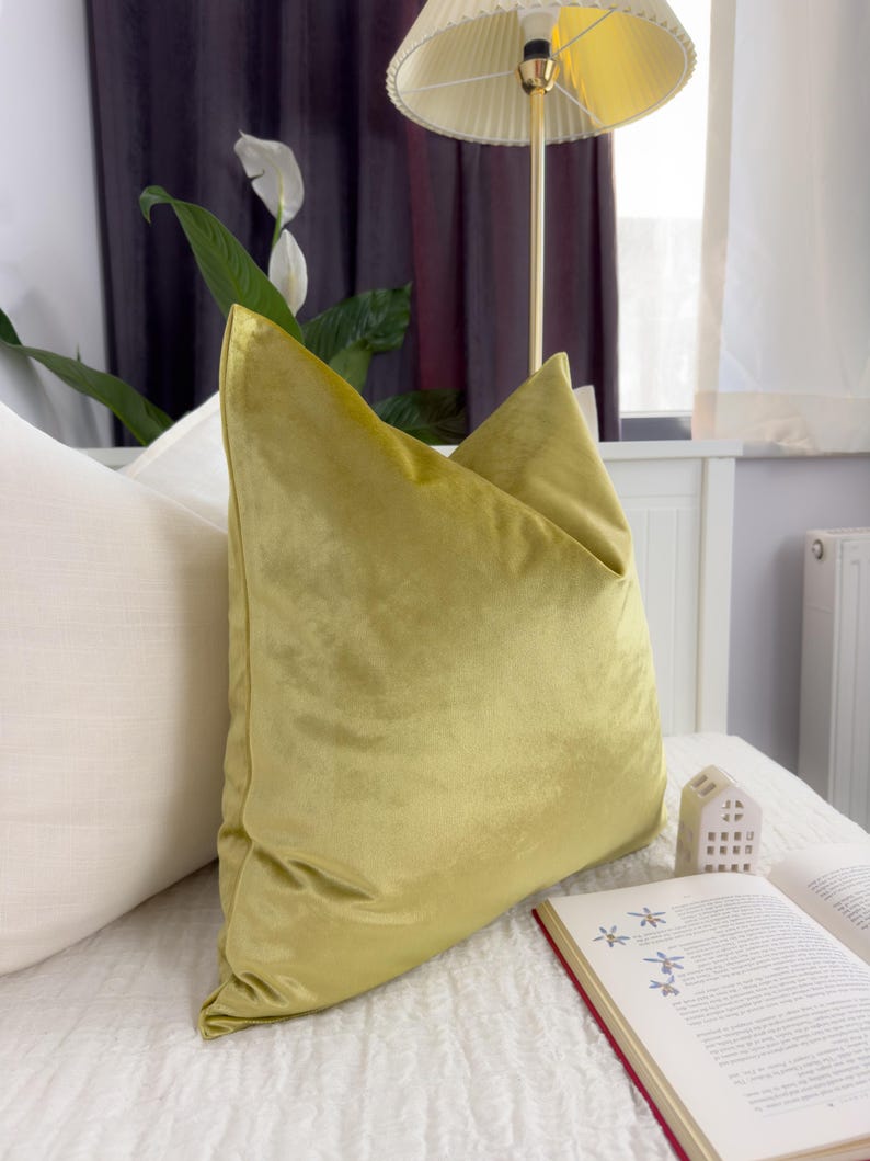 K&ouml;nnte beinhalten: Ein kissenf&ouml;rmiges Dekokissen aus chartreusefarbenem Samt liegt auf einem Bett neben einem wei&szlig;en Kissen. Das Kissen hat ein weiches, pl&uuml;schiges Aussehen. Ein Buch mit rotem Buchr&uuml;cken und ein kleines dekoratives Haus liegen ebenfalls auf dem Bett.