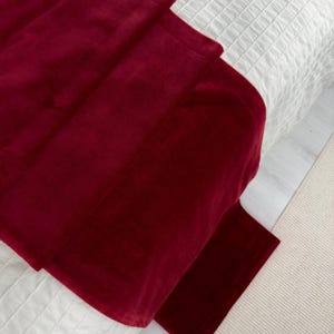 May include: A burgundy velvet throw blanket folded on a white bedspread. The blanket is folded in thirds and the edges are visible.