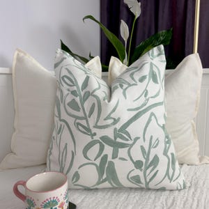 May include: A decorative pillow with a white background and a green leaf pattern. Two solid white pillows flank the patterned pillow. A floral mug with a pink handle sits on a black coaster. The scene is set on a white bed.
