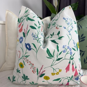 May include: White throw pillow with a colorful floral pattern featuring pink, blue, yellow, and green flowers and leaves. The pillow is on a white bed with a book and a cup in the foreground. A second pillow with a green leaf pattern is visible.