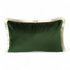 Puede incluir: Un cojín rectangular de terciopelo verde oscuro con un borde de flecos color crema. El cojín es de color liso y tiene un aspecto suave y afelpado. La franja añade un elemento decorativo a los bordes del cojín.
