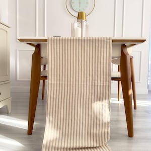 May include: A beige and brown striped table runner with a subtle linen texture. The runner is draped over a white table with wooden legs and is positioned in front of two chairs.