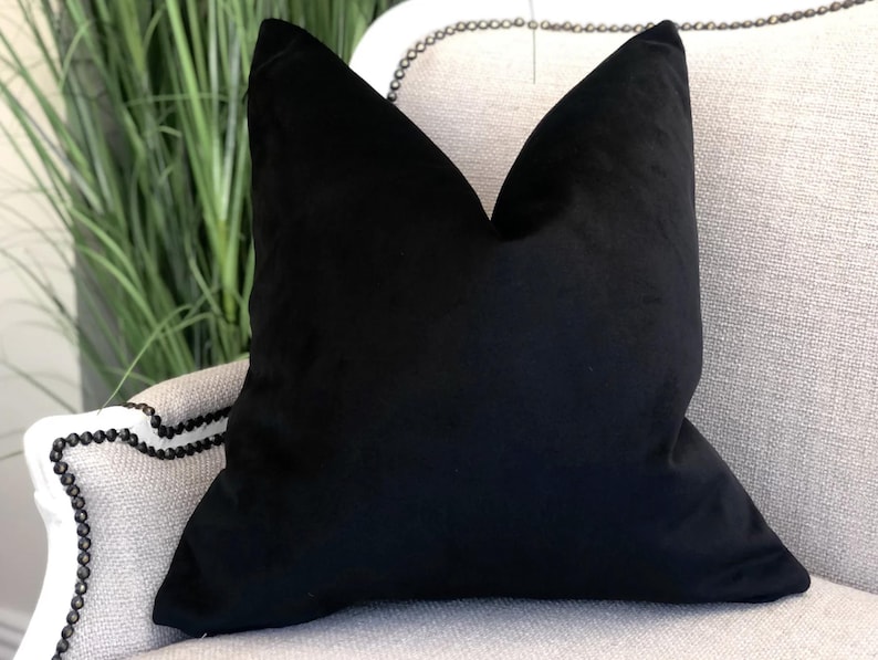 May include: A black velvet cushion with a square shape and a smooth texture. The cushion is sitting on a light grey sofa with a beaded trim.