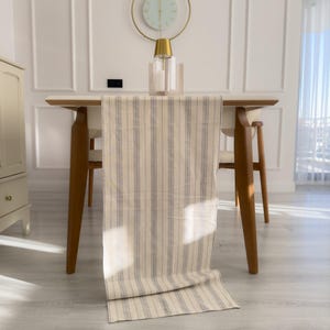 Puede incluir: Un camino de mesa a rayas beige y grises sobre una mesa de madera. La mesa tiene patas de madera y está en una habitación luminosa. Un reloj dorado y un candelabro están sobre la mesa.