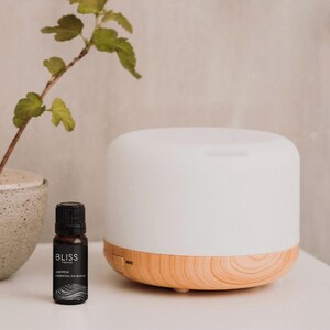 May include: A white essential oil diffuser with a wood grain base is on a white surface. A small bottle of essential oil and a potted plant with green leaves are also present. The bottle has the text "BLISS" and "centre essential oil blend".