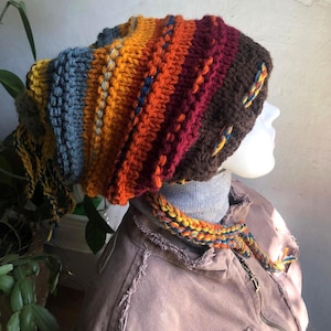 Puede incluir: Un gorro de punto grueso con rayas horizontales en tonos marrón, burdeos, naranja, amarillo y azul. El gorro presenta un patrón de ganchillo texturizado con borlas de hilo trenzado colgantes a los lados y un detalle de pompón detrás.