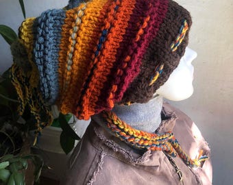 May include: A chunky knit beanie featuring horizontal stripes in shades of brown, burgundy, orange, yellow, and blue. The hat has a textured crochet pattern with dangling braided yarn tassels on the sides and a pom-pom detail at the back.