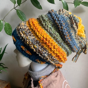 May include: A handmade, colorful knit beanie in shades of blue, orange, green, and yellow. The hat has a textured, chunky knit design and a long, slouchy fit. The beanie is a stylish accessory for cold weather.