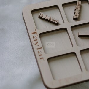 May include: A wooden tray with nine square compartments, each with a cutout for a small wooden tile. The tray is labeled "Taylor Couture" and has the phrase "He Is Risen" written on two of the tiles.