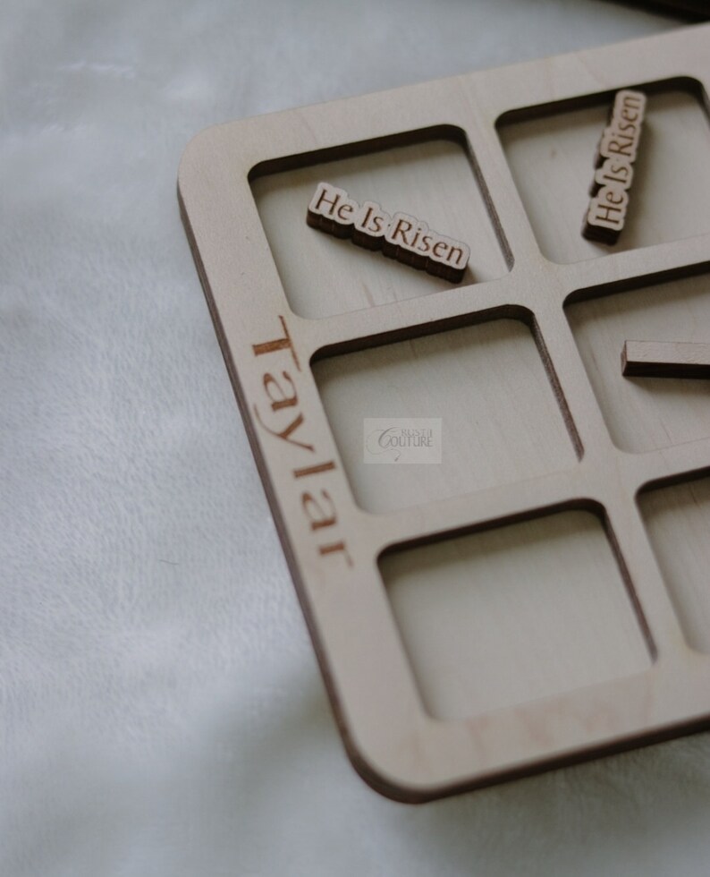 May include: A wooden tray with nine square compartments, each with a cutout for a small wooden tile. The tray is labeled "Taylor Couture" and has the phrase "He Is Risen" written on two of the tiles.