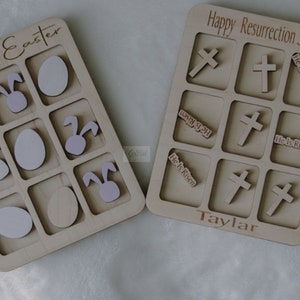 May include: Two wooden game boards with a tic-tac-toe style grid. The board on the left says "Happy Easter" and has bunny and egg images. The board on the right says "Happy Resurrection Sunday" and has crosses and "He is Risen" text.