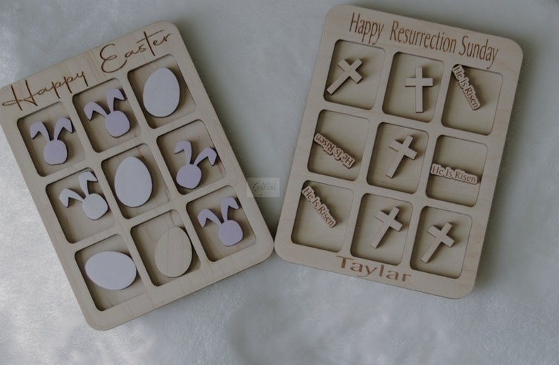 May include: Two wooden game boards with a tic-tac-toe style grid. The board on the left says "Happy Easter" and has bunny and egg images. The board on the right says "Happy Resurrection Sunday" and has crosses and "He is Risen" text.