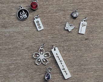 May include: A collection of silver-toned metal charms including a circular flame pendant, a red crystal, a rectangular tag with three circles, a small heart with a clear stone, a butterfly, a flower, a long bar with cursive text, and a teardrop charm.