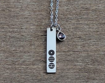 Op de afbeelding: Een zilverkleurige ketting met een verticale rechthoekige hanger. De hanger heeft het woord "ODE" in een gestileerd lettertype en een kleine paarse traanvormige bedel. De ketting wordt getoond op een houten oppervlak.