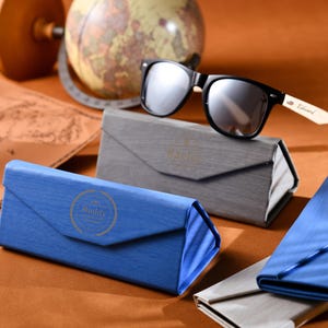 Puede incluir: Dos estuches plegables para gafas de sol azules y uno gris con un par de gafas de sol negras con patillas de madera. El estuche para gafas de sol tiene la palabra "Buddy" impresa.