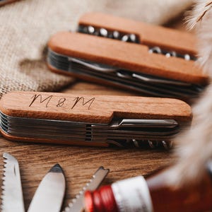 Peut inclure: Deux couteaux de poche &agrave; manche en bois avec plusieurs outils. Le couteau du haut a un manche brun et le couteau du bas a un manche brun clair. Le couteau du bas porte les initiales "M&M" grav&eacute;es sur le manche.