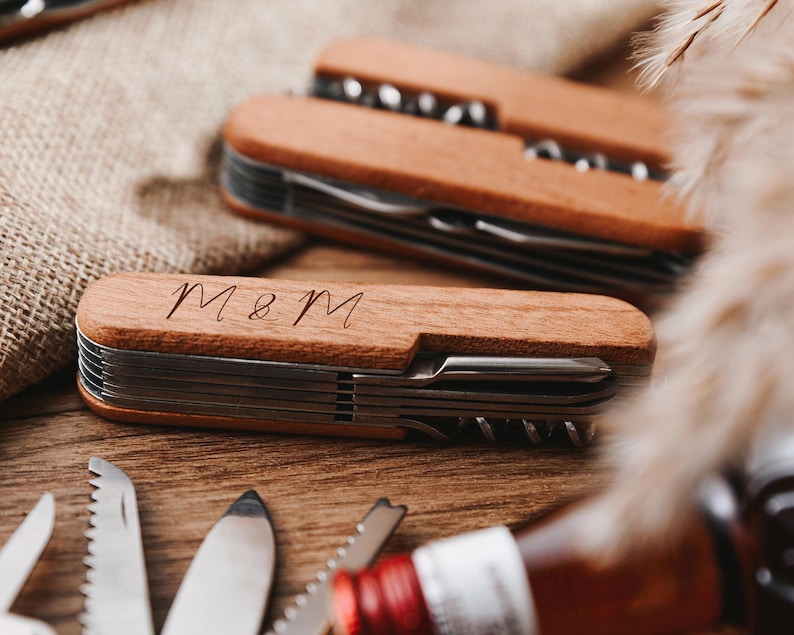 Peut inclure: Deux couteaux de poche &agrave; manche en bois avec plusieurs outils. Le couteau du haut a un manche brun et le couteau du bas a un manche brun clair. Le couteau du bas porte les initiales "M&M" grav&eacute;es sur le manche.