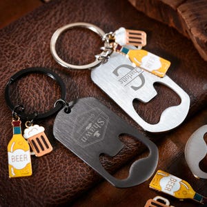 Peut inclure: Deux porte-clés ouvre-bouteille en argent avec du texte gravé. Un porte-clés porte le texte "Charlotte" et l'autre porte le texte "Shawn Established 1984". Les deux porte-clés ont un charme de bouteille de bière et de chope à bière.